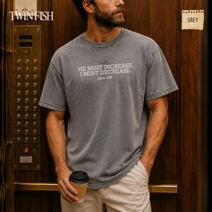 May include: A grey short-sleeved t-shirt featuring the white printed text HE MUST INCREASE, I MUST DECREASE, John 3:30. The shirt has a crew neckline and a relaxed fit, worn with light-colored trousers and a thin silver chain necklace.