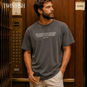 May include: A charcoal gray short-sleeve t-shirt featuring the white printed text "HE MUST INCREASE, I MUST DECREASE. John 3:30" across the chest. The shirt has a crew neckline and a relaxed fit, paired with light-colored pants and a thin silver chain necklace.
