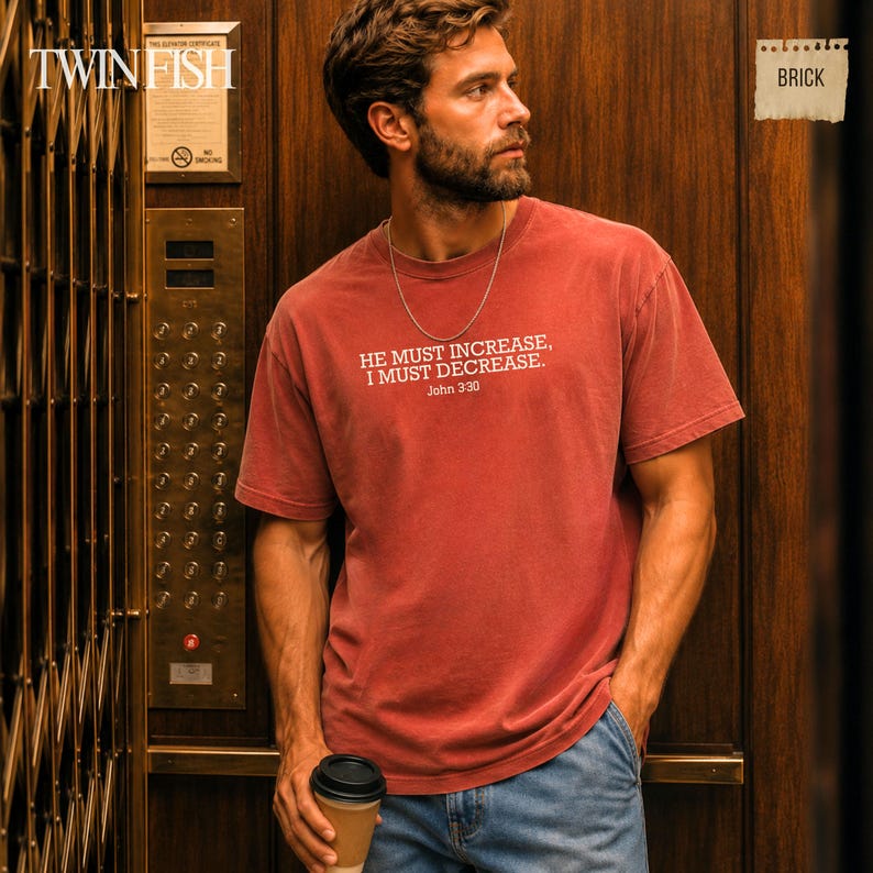 May include: A brick-colored short-sleeve t-shirt featuring the white printed text HE MUST INCREASE, I MUST DECREASE. John 3:30. The shirt has a crew neckline and is worn with a thin silver chain necklace and blue denim jeans.