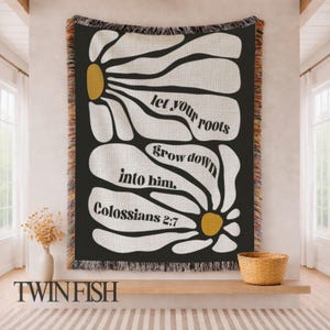 May include: A woven throw blanket with a black background and a large white daisy design. The text "let your roots grow down into him. Colossians 2:7" is displayed in white. The blanket has a fringed edge.