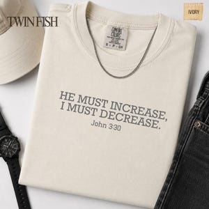 May include: An ivory t-shirt featuring the dark gray text HE MUST INCREASE, I MUST DECREASE. John 3:30 printed on the chest. The shirt has a Comfort Colors label at the neck, a silver chain necklace, and is displayed with a cap and a watch.