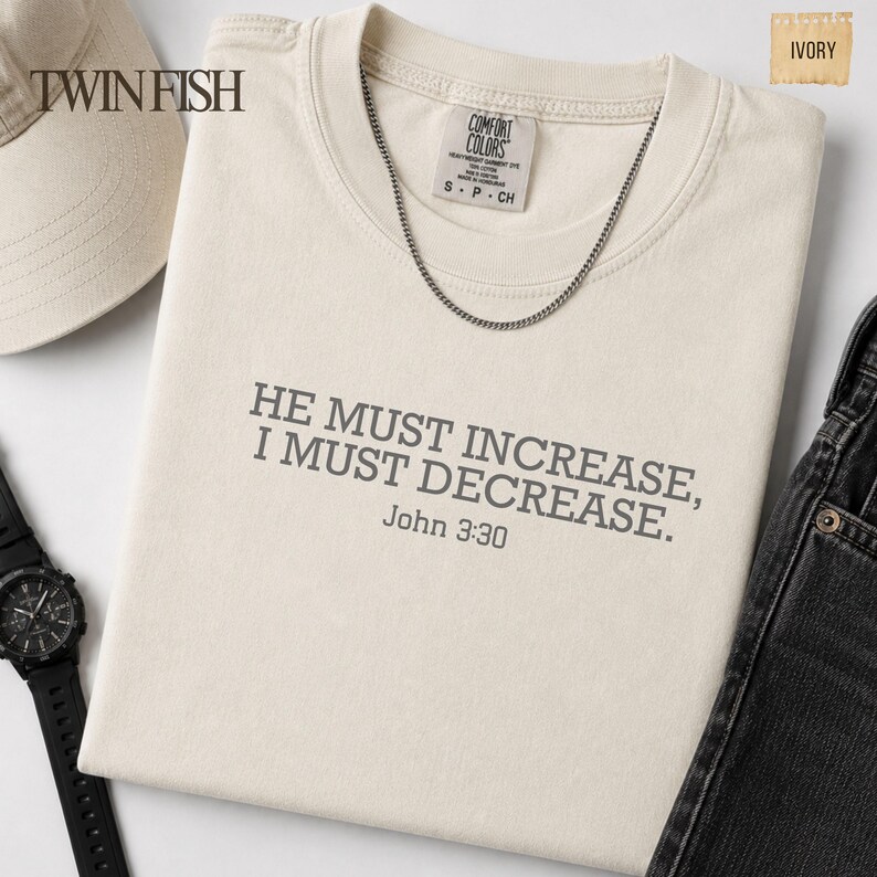 May include: An ivory t-shirt featuring the dark gray text HE MUST INCREASE, I MUST DECREASE. John 3:30 printed on the chest. The shirt has a Comfort Colors label at the neck, a silver chain necklace, and is displayed with a cap and a watch.