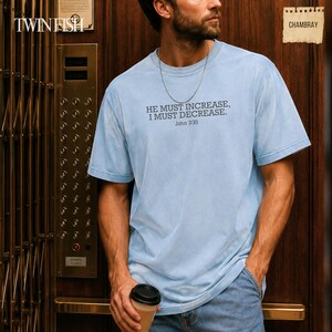 May include: A light blue short-sleeve t-shirt featuring the dark grey text HE MUST INCREASE, I MUST DECREASE. John 3:30 printed on the chest. The shirt has a crew neckline and a relaxed fit, paired with a thin silver chain necklace.