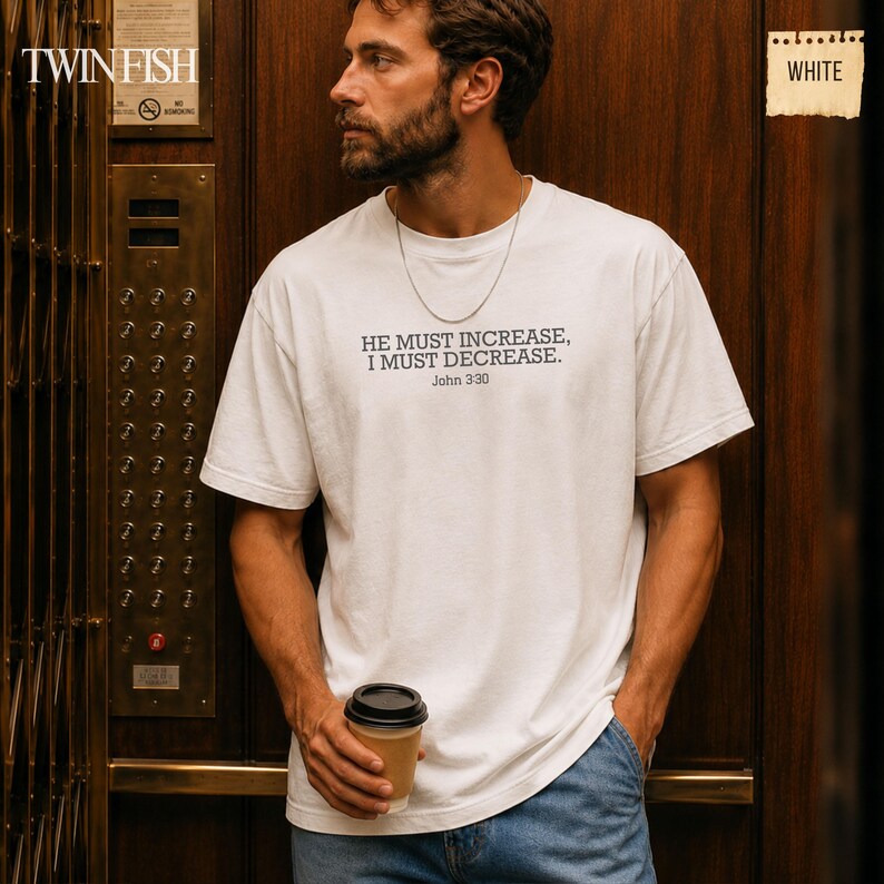 May include: A white short-sleeved t-shirt featuring the black printed text HE MUST INCREASE, I MUST DECREASE. John 3:30 centered on the chest. The garment is worn with a thin silver chain necklace and blue denim jeans, paired with a coffee cup.