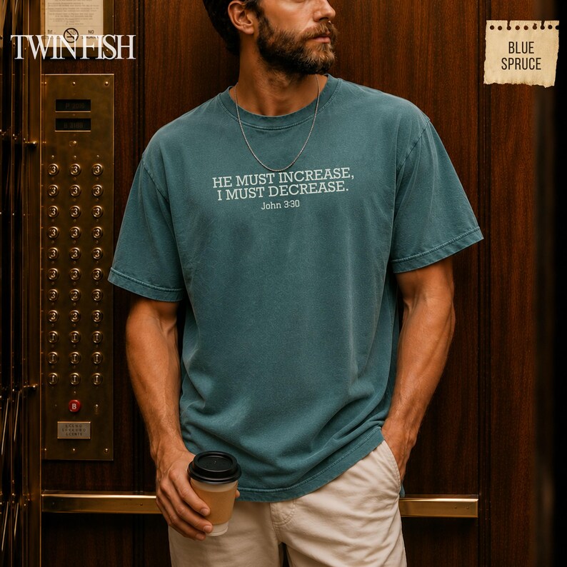 May include: A blue spruce colored short-sleeve t-shirt featuring the white printed text HE MUST INCREASE, I MUST DECREASE. John 3:30 centered on the chest. The garment has a crew neckline and a relaxed fit, paired with light-colored trousers.