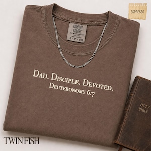 Op de afbeelding: Een espresso-kleurig t-shirt met korte mouwen en de witte tekst "DAD. DISCIPLE. DEVOTED. DEUTERONOMY 6:7" op de borst gedrukt. Het shirt bevat een zilverkleurige ketting die op de kraag rust en een label van het merk Comfort Colors.
