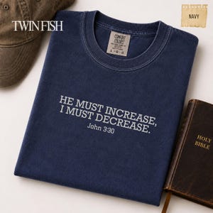 May include: A navy blue crew neck t-shirt featuring the white embroidered text HE MUST INCREASE, I MUST DECREASE. John 3:30. The shirt has a Comfort Colors label inside the collar, with the size marked as L G G, and is displayed alongside a brown cap and a book.