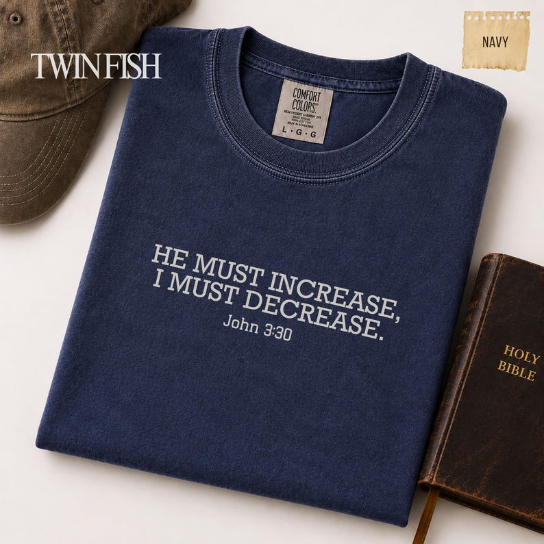 May include: A navy blue crew neck t-shirt featuring the white embroidered text HE MUST INCREASE, I MUST DECREASE. John 3:30. The shirt has a Comfort Colors label inside the collar, with the size marked as L G G, and is displayed alongside a brown cap and a book.
