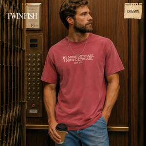 May include: A crimson short-sleeve t-shirt featuring the white text HE MUST INCREASE, I MUST DECREASE. John 3:30 printed on the chest. The shirt has a crew neckline and a relaxed fit, paired with blue denim jeans and a silver chain necklace.