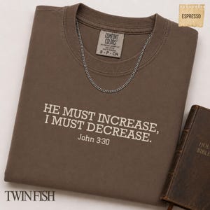 May include: A brown short-sleeved t-shirt featuring the white text HE MUST INCREASE, I MUST DECREASE. John 3:30 printed on the chest. The shirt has a Comfort Colors label at the neck and is displayed with a silver chain necklace and a brown book.