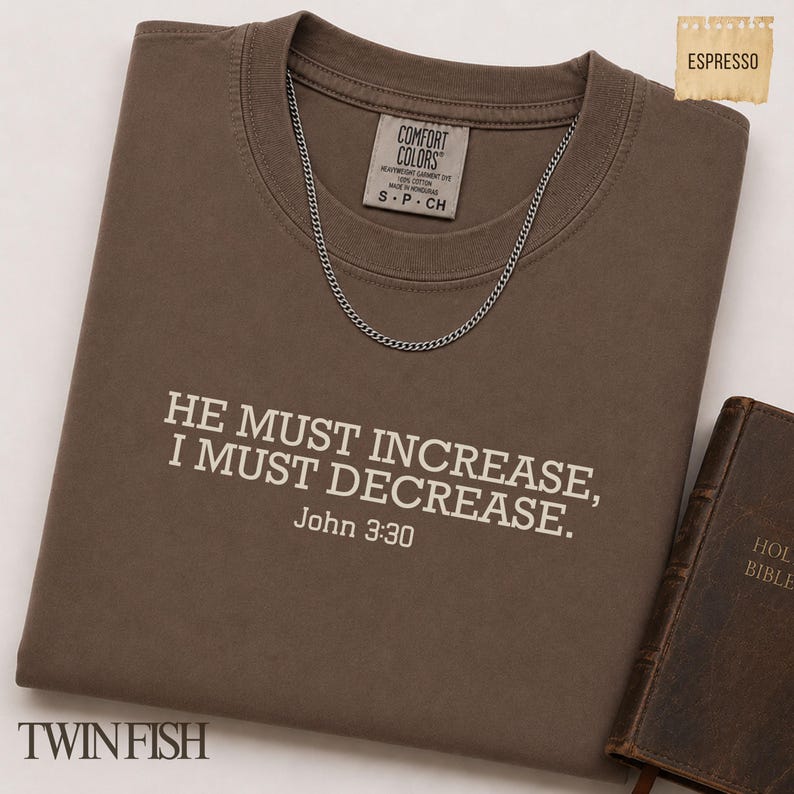 May include: A brown short-sleeved t-shirt featuring the white text HE MUST INCREASE, I MUST DECREASE. John 3:30 printed on the chest. The shirt has a Comfort Colors label at the neck and is displayed with a silver chain necklace and a brown book.