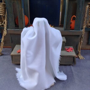 May include: A white ghost costume with a draped fabric design. The ghost is standing in front of a miniature Halloween scene with a blue door, a pumpkin, and a skeleton. The costume has a frayed edge.