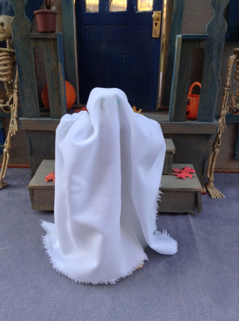 May include: A white ghost costume with a draped fabric design. The ghost is standing in front of a miniature Halloween scene with a blue door, a pumpkin, and a skeleton. The costume has a frayed edge.