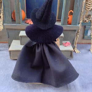May include: A black witch doll wearing a black pointed hat and a black cape. The doll is standing on a wooden step with a red leaf on the ground.