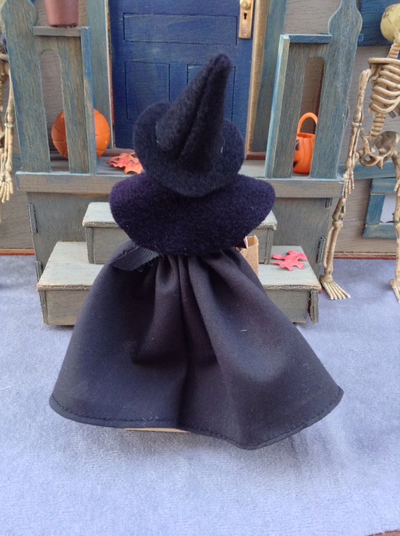 May include: A black witch doll wearing a black pointed hat and a black cape. The doll is standing on a wooden step with a red leaf on the ground.
