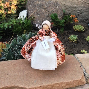 May include: A handmade doll wearing a red, brown, and white patterned dress with a white apron. The doll has a wooden head with painted features and a tag that reads "Prairie".