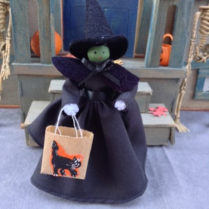 May include: A witch doll wearing a black dress and hat, holding a brown paper bag with a black cat illustration. The doll has green skin and black eyes.