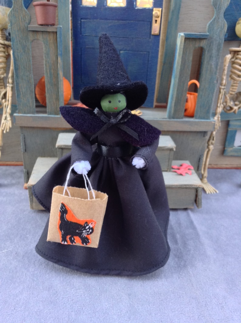 May include: A witch doll wearing a black dress and hat, holding a brown paper bag with a black cat illustration. The doll has green skin and black eyes.