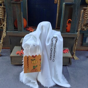 May include: A miniature Halloween scene featuring a white ghost figure with a decorative chain and a small orange and brown trick-or-treat bag. The bag is decorated with pumpkins and other fall motifs. The scene is set on a porch with a blue door.