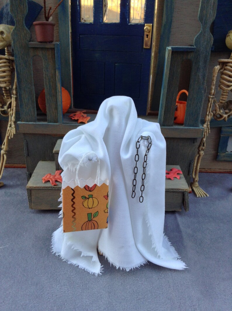 May include: A miniature Halloween scene featuring a white ghost figure with a decorative chain and a small orange and brown trick-or-treat bag. The bag is decorated with pumpkins and other fall motifs. The scene is set on a porch with a blue door.