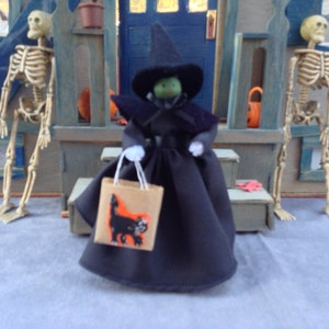 May include: A witch doll wearing a black dress and a pointed black hat with a green face. The doll is holding a brown paper bag with a black cat illustration. The doll is standing in front of a wooden door with two skeletons on either side.