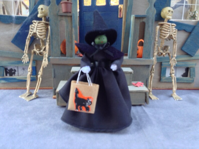 May include: A witch doll wearing a black dress and a pointed black hat with a green face. The doll is holding a brown paper bag with a black cat illustration. The doll is standing in front of a wooden door with two skeletons on either side.