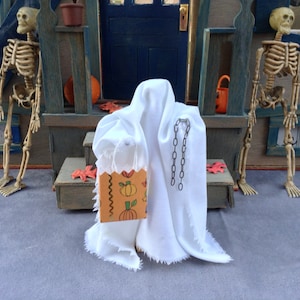 May include: A miniature Halloween scene featuring a white ghost figure holding a trick-or-treat bag with pumpkin illustrations. Two skeleton figures stand on either side of a miniature porch. The scene is set against a blue and gray backdrop.