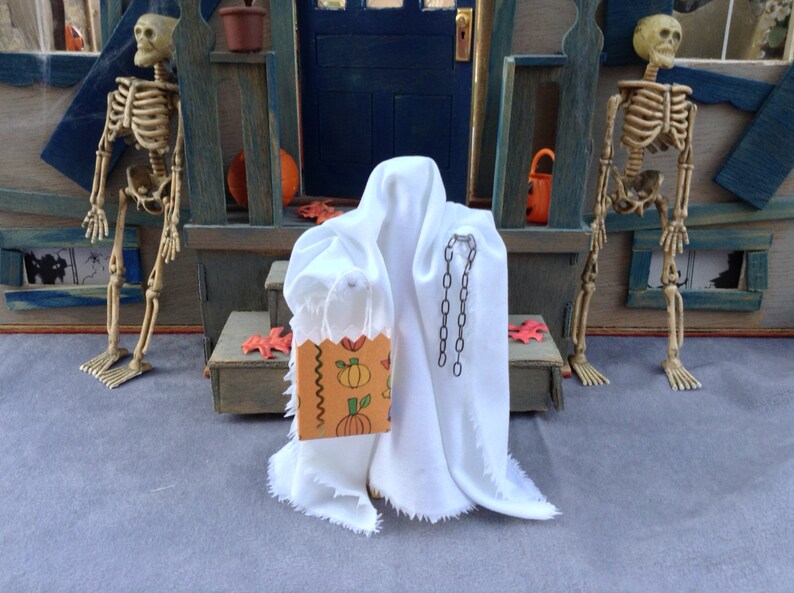 May include: A miniature Halloween scene featuring a white ghost figure holding a trick-or-treat bag with pumpkin illustrations. Two skeleton figures stand on either side of a miniature porch. The scene is set against a blue and gray backdrop.