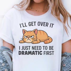 Puede incluir: Camiseta blanca con el texto "I'll Get Over It, I Just Need To Be Dramatic First". La camiseta presenta una ilustración de dibujos animados de un gato atigrado naranja con una expresión gruñona.