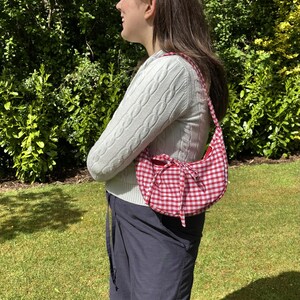 May include: A red and white gingham pattern shoulder bag with a matching headband. The bag has a curved shape and a bow detail. The person is wearing a white cable knit sweater and navy blue cargo pants.