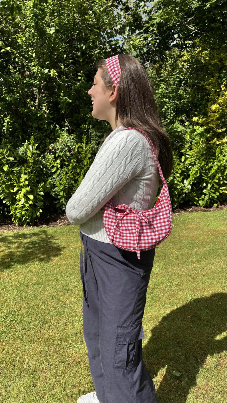 May include: A red and white gingham pattern shoulder bag with a matching headband. The bag has a curved shape and a bow detail. The person is wearing a white cable knit sweater and navy blue cargo pants.