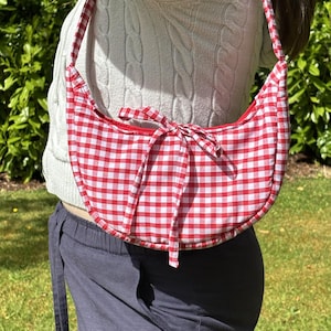 May include: Red and white gingham pattern crescent-shaped shoulder bag with a matching strap and bow closure. The bag is made of fabric and has a red interior. The bag is worn over the shoulder.