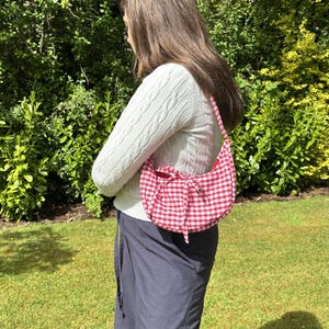 May include: Red and white gingham pattern shoulder bag with a bow detail. The bag is crescent-shaped and has a matching strap. The background includes green foliage and grass.