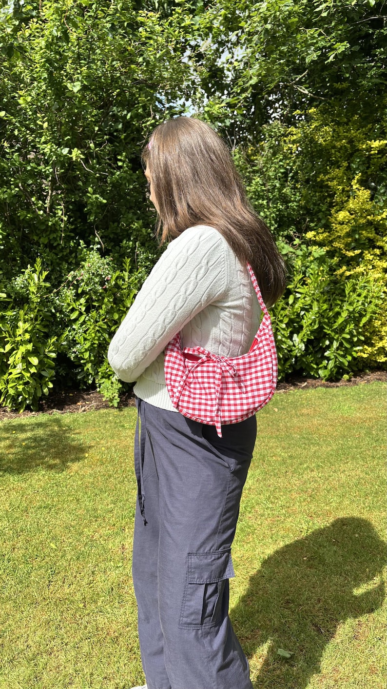 May include: Red and white gingham pattern shoulder bag with a bow detail. The bag is crescent-shaped and has a matching strap. The background includes green foliage and grass.