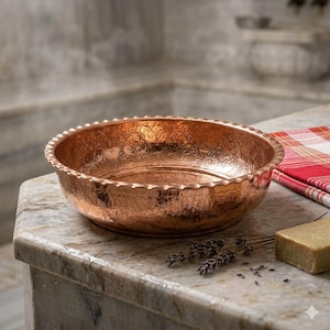 Puede incluir: Un cuenco de cobre martillado con un patrón floral decorativo y un borde festoneado. El cuenco es de color cobre cálido y está sobre una superficie de mármol. Una tela a cuadros roja y blanca y jabón están cerca.