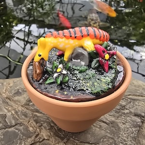 May include: A small terracotta pot containing a sculpted scene with a yellow and orange striped creature, a grey stone, green foliage, and red flowers. The creature has a long neck and is positioned over a brown rock within the textured base.
