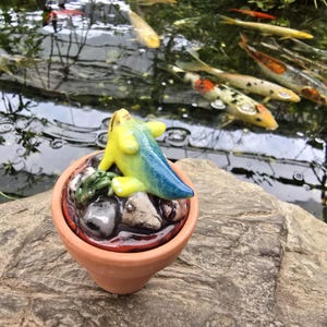 May include: A small terracotta pot containing a ceramic sculpture of a yellow and blue creature resting on a cluster of smooth gray and white stones. The sculpture features a smooth, glossy finish and is positioned outdoors near a pond with koi fish.