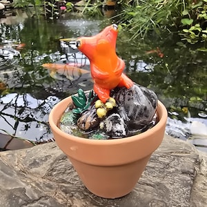 May include: A bright orange glass frog figurine perched on a cluster of dark gray rocks and small green accents, all contained within a simple terracotta-colored clay pot. The frog has prominent eyes and a smooth, glossy finish on its surface.