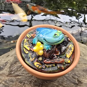 May include: A small terracotta bowl containing a sculpted scene of a bright blue frog resting on a dark, textured base. The base features yellow mushroom-like clusters, brown twigs, and scattered yellow leaves, all finished with a glossy, wet-look coating.