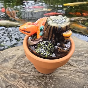 May include: A small terracotta pot containing a sculpted orange salamander resting on a dark brown tree stump. The stump features textured bark details and a patch of green moss. The salamander has a smooth, bright orange body and a distinct head.