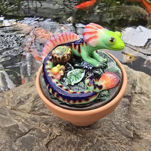 May include: A small terracotta pot containing a sculpted lizard figurine with a green body, a frilled head, and a long, curved tail featuring blue, orange, and purple accents. The lizard rests on a base with small sculpted leaves and a tree stump.