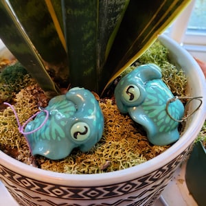 May include: Two turquoise ceramic ornaments shaped like stylized creatures with large eyes and patterned details. Each ornament has a loop for hanging, one with a purple string and the other with a green string. They rest on moss inside a decorative pot.
