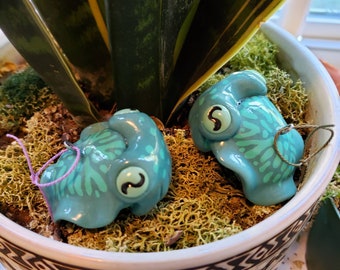 May include: Two turquoise ceramic ornaments shaped like stylized creatures with large eyes and patterned details. Each ornament has a loop for hanging, one with a purple string and the other with a green string. They rest on moss inside a decorative pot.