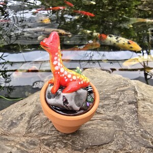 May include: A small, bright orange dinosaur figurine with white spots, perched atop a smooth gray stone inside a miniature terracotta pot. The dinosaur has a long neck and a rounded head, and the stone base is nestled within the clay container.