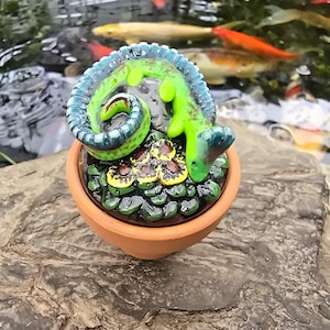 May include: A small terracotta pot containing a sculpted green lizard with a curled tail resting on a cluster of dark green leaves and yellow flowers. The lizard features blue accents along its back and tail, with a smooth, glossy finish throughout.
