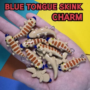 May include: A collection of nine blue-tongued skink charms made of polymer clay. The charms are painted with brown and orange stripes and have blue accents on the tongue and eyes. The charms are displayed on a hand, with the text "BLUE TONGUE SKINK CHARM" visible in the background.