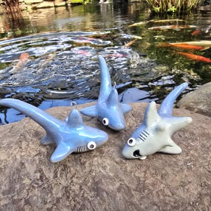 May include: Three ceramic shark figurines in shades of blue and gray are displayed on a rock. The sharks have detailed features, including fins, eyes, and open mouths. The background shows a pond with koi fish.