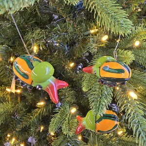 May include: Three colorful Christmas ornaments shaped like stylized bugs, hanging on a Christmas tree. The ornaments are primarily orange and green with red accents. The tree is a lush green with small white lights.