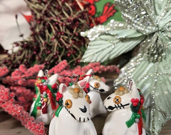 May include: Four white ceramic horse ornaments with black stitched facial features and gold accents. Each horse has a red and green garland and a silver loop for hanging. Festive holiday decor.