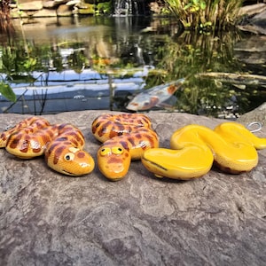 Puede incluir: Una colección de figuras de serpientes de cerámica en varios tamaños y poses. Las serpientes presentan un patrón de rayas amarillas y marrones, algunas con rasgos faciales de dibujos animados. Las figuras se exhiben sobre una superficie de piedra gris texturizada.